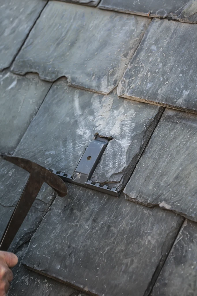 Close-up of a SlatePlate metal mounting bracket installed flush between gray slate tiles, with a slate ripping tool alongside.