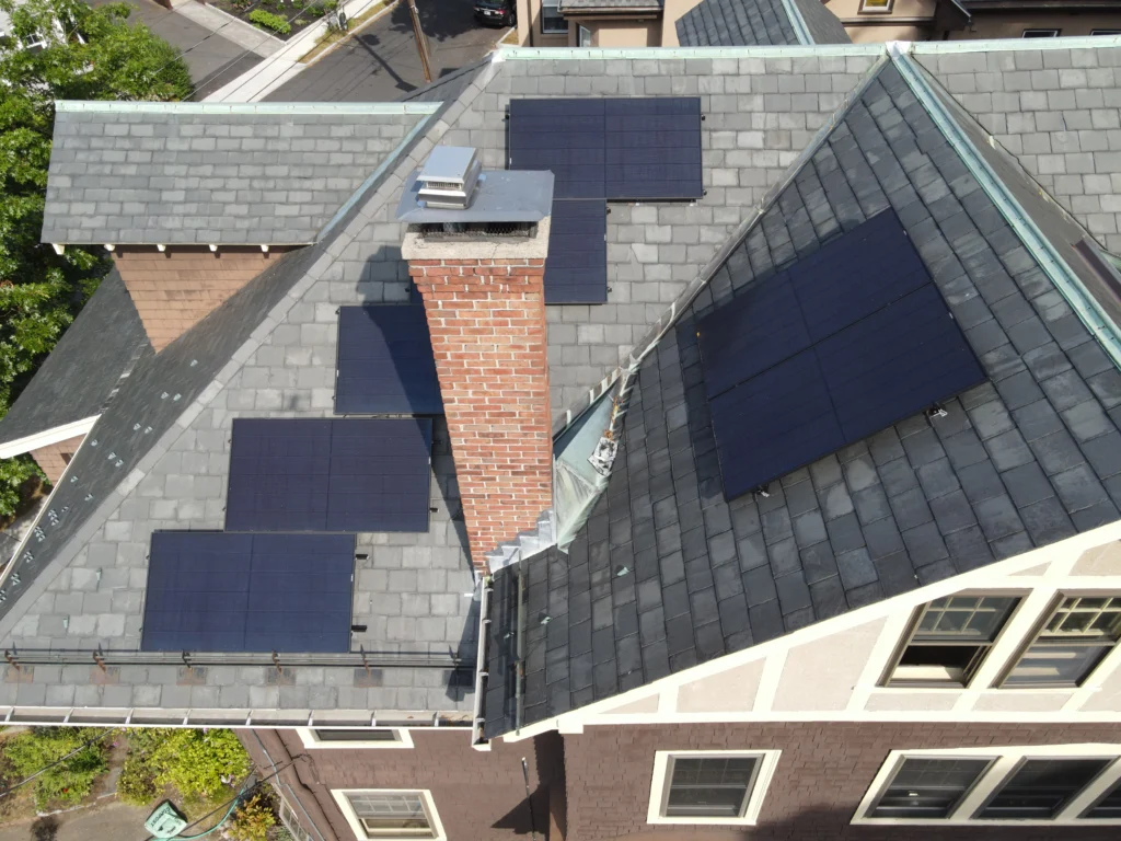 Aerial view of black solar sub-arrays mounted around a central brick chimney on the gray slate roof of a home in Watertown, Massachusetts.
